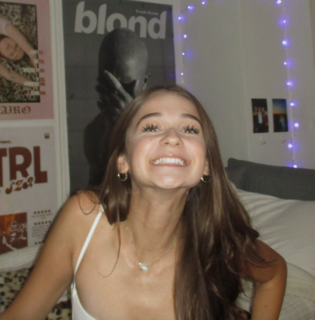 Bea Lloyd wears an expression of joy while sitting on a bed in front of a Frank Ocean poster.